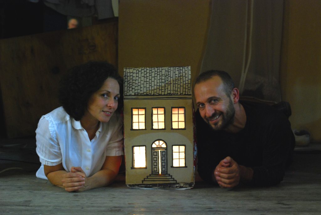 I due protagonisti sono sdraiati per terra a pancia in giù, tirati su con la schiena appoggiandosi sugli avambracci. Guardano in camera sorridendo. In mezzo a loro, una piccola casa di cartone, illuminata all'interno, alta fino alle loro teste.