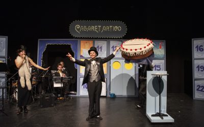 Tutti gli attori e musicisti sul palco in un grandangolo, mentre allargano le braccia verso il pubblico. Uno di loro sorregge un'enorme torta fatta di stoffa