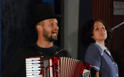 Primo piano dei due protagonisti. A sinistra, lui suona la fisarmonica e canta, con un cilindro in testa. Lei, a destra, balla.