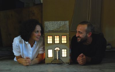 I due protagonisti sono sdraiati per terra a pancia in giù, tirati su con la schiena appoggiandosi sugli avambracci. Si guardano sorridendo. In mezzo a loro, una piccola casa di cartone, illuminata all'interno, alta fino alle loro teste.