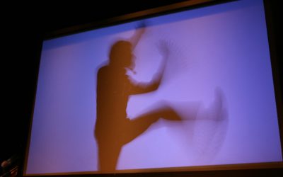 Su un telo è stato proiettato uno sfondo viola. Dietro il telo si trova un attore, la cui ombra si riflette su questo. La foto è stata scattata con tempi lunghi e quindi si vede la scia del movimento della gamba sollevata e delle braccia dell'ombra dell'attore.