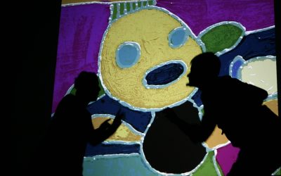 Un'immagine di un disegno colorato raffigurante una persona stilizzata è proiettata su un telo sul palco. Sull'immagine possiamo vedere le ombre dei due attori che si trovano dietro il telo.