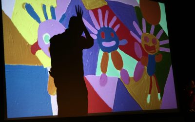 Un'immagine di un disegno colorato raffigurante tre persone stilizzate è proiettata su un telo sul palco. Sull'immagine possiamo vedere l'ombra di un attore che si trova dietro il telo. Ha una mano alzata sopra la testa con le dita larghe, facendo quasi una cresta, imitando i disegni.