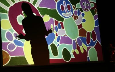 Un'immagine di un disegno colorato raffigurante tre persone stilizzate è proiettata su un telo sul palco. Sull'immagine possiamo vedere l'ombra di un attore che si trova dietro il telo. Ha le braccia vicino al busto che si aprono, con le dita larghe, stile jazz hands, imitando i disegni.