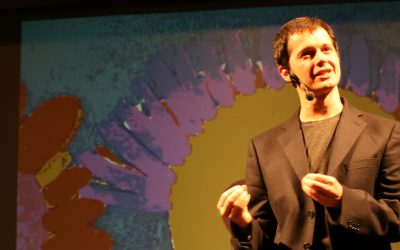 Piano americano dell'attore sul palco. Indossa una camicia nera e sta gesticolando. Dietro di lui, un disegno proiettato, coloratissimo, raffigurante un grosso sole.