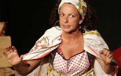 Primo piano della protagonista, che parla col pubblico. Indossa una fascia nei capelli fatta con panni da cucina, che ha anche sulle spalle a scialle. Indossa un grembiule da cucina e delle tovaglie come gonna.