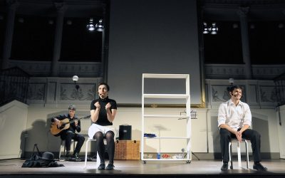 Un attore e un'attrice sul palco durante lo spettacolo, seduti su degli sgabelli. Dietro di loro un musicista suona la chitarra.