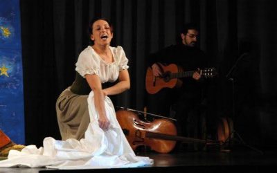La lavandaia, in ginocchio, raccoglie le fasce da terra, mentre un musicista, dietro di lei, suona la chitarra.